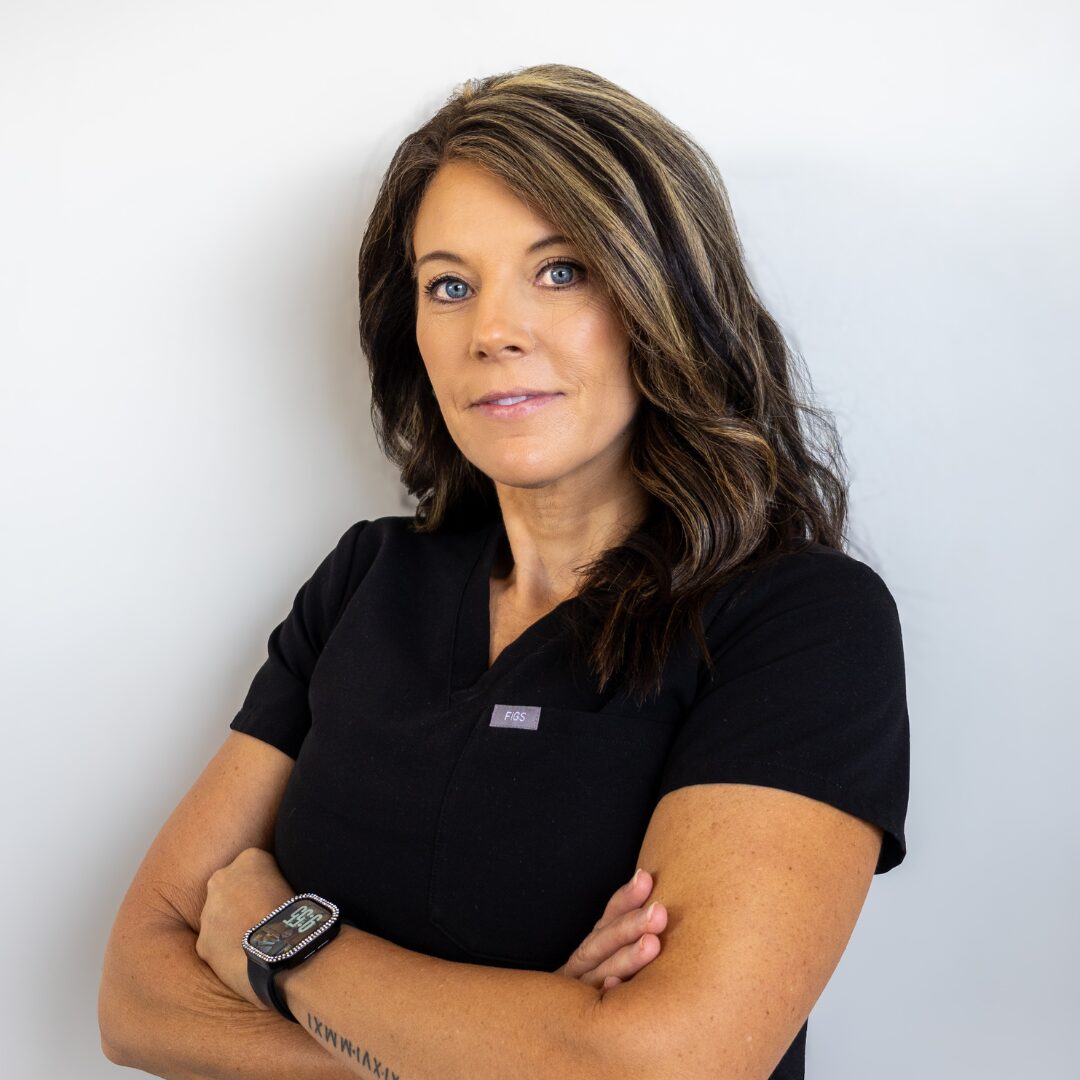 Kim Ellis of Silique Companies in black scrubs with arms crossed, smiling softly against a light gray background.