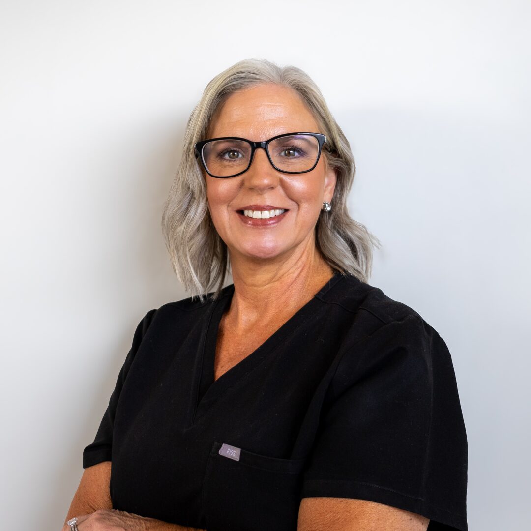 Kim Nix of Silique Companies smiling in black scrubs and glasses, with silver-gray hair, against a light gray background.